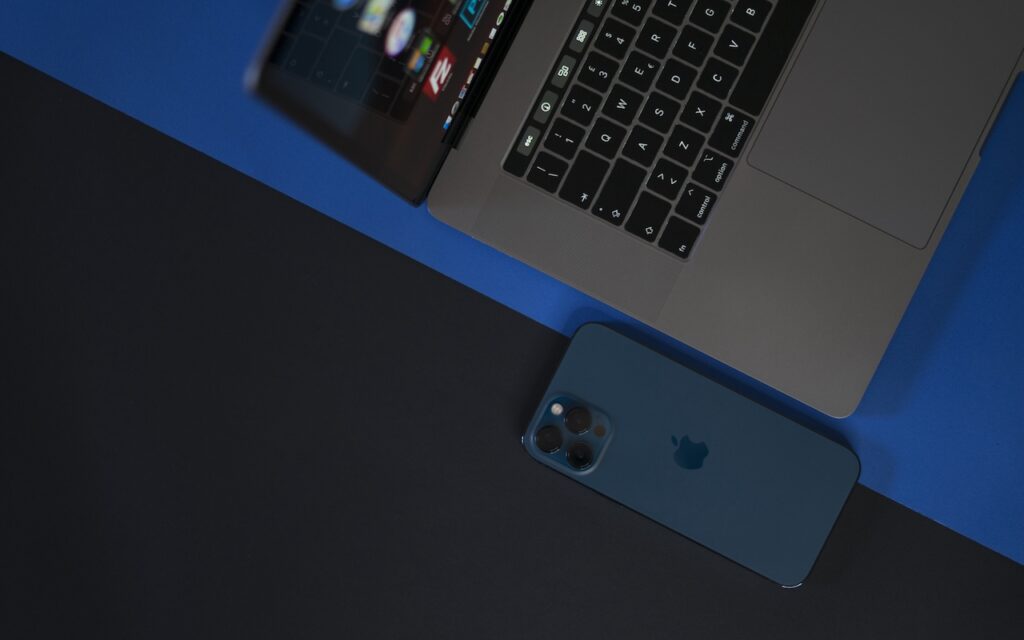 A blue iPhone lies next to the keyboard of an open silver MacBook laptop on a blue and black surface, hinting at How (and Why) to Use iPhone Mirroring on Your Mac. The devices are seen from above, with the phone partially under the laptop.