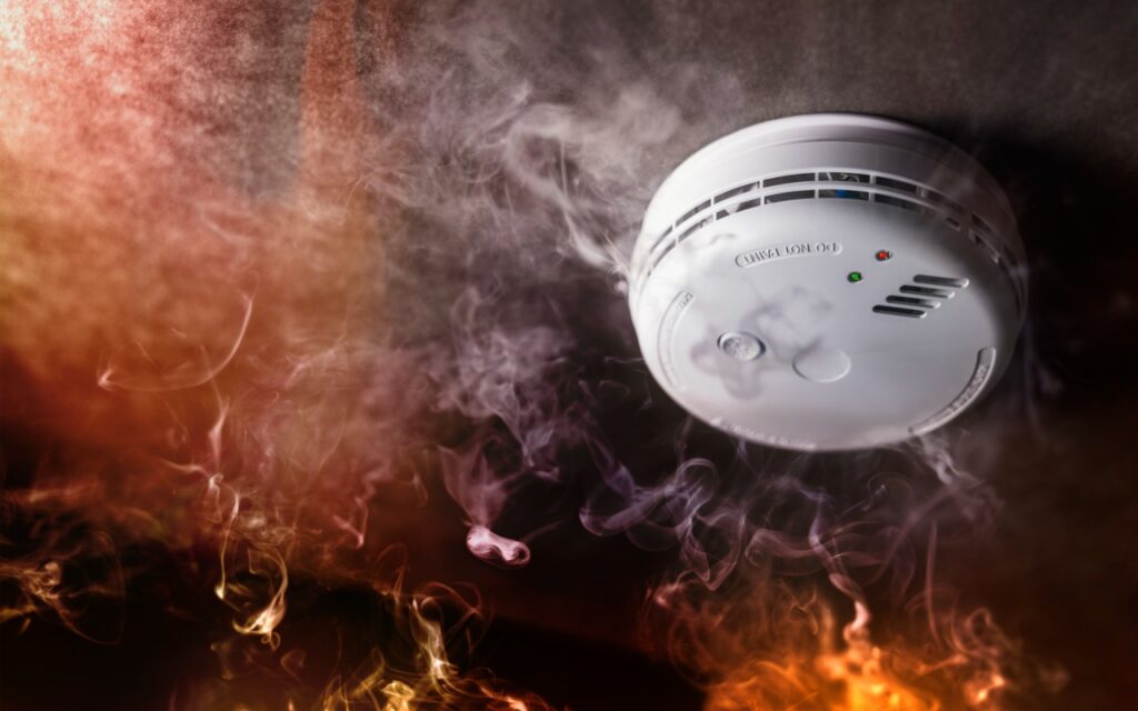 A ceiling-mounted smoke detector surrounded by smoke and flames, with indicator lights visible on the device—illustrating a fire emergency, where HomePods can alert you of smoke and carbon monoxide alarms for added safety.