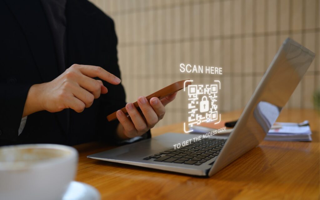 A person holds a smartphone and points at a glowing wifi QR code projected above a laptop, with “SCAN HERE TO GET THE ACCESS CODE” displayed, making it easy to share Wi-Fi network passwords using QR codes. A coffee cup sits on the wooden table in the foreground.