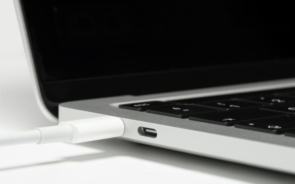 Close-up of a white USB-C charging cable plugged into the side of a silver laptop with a black keyboard. The screen is partially visible and turned off—a reminder that Apple Silicon Macs can’t boot from the DFU port.