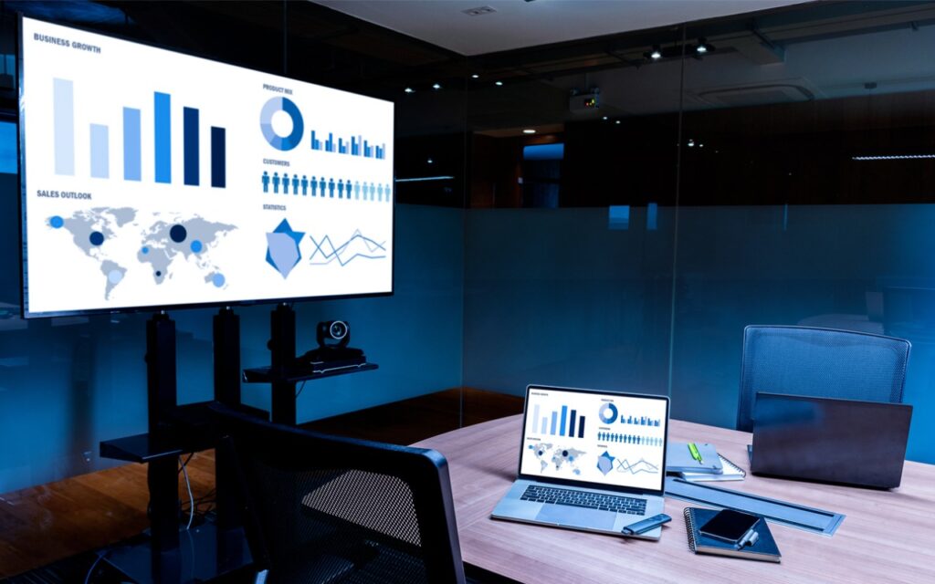 A modern office meeting room with a large screen displaying business charts and graphs, a laptop showing similar data—ready for you to use AirPlay to mirror or extend your Mac’s display—plus a tablet, smartphone, and an empty chair at the conference table.