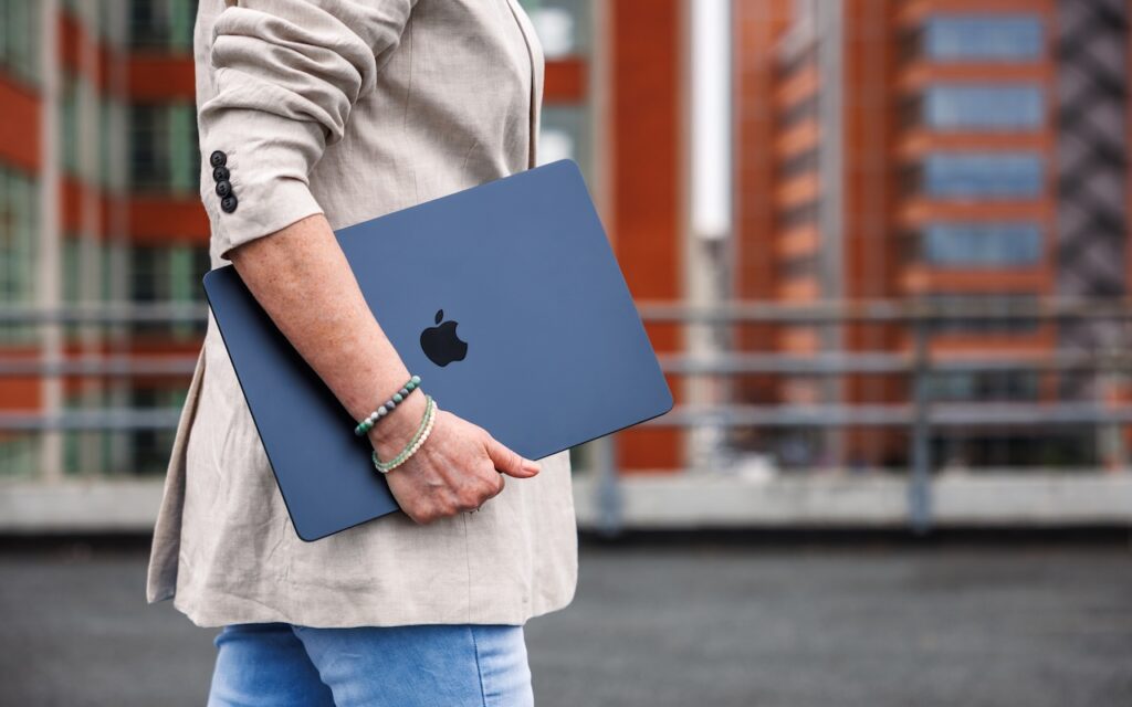 A person in a beige blazer and blue jeans holds a closed, dark-colored Apple MacBook under their arm—perhaps considering Choosing the Best Mac for a College-Bound Student in 2025—while standing outdoors with blurred buildings in the background.