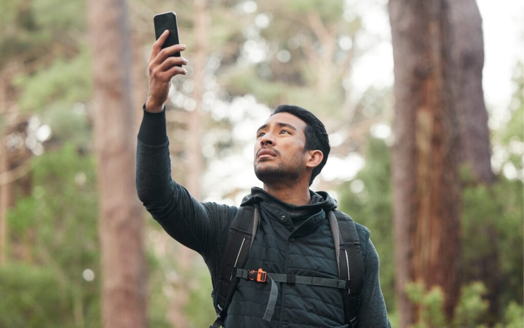 A person wearing a backpack holds a smartphone up in the air while standing in a forest, trying to stay connected off the grid—perhaps using Messages via Satellite in iOS 18—while searching for signal among tall trees.