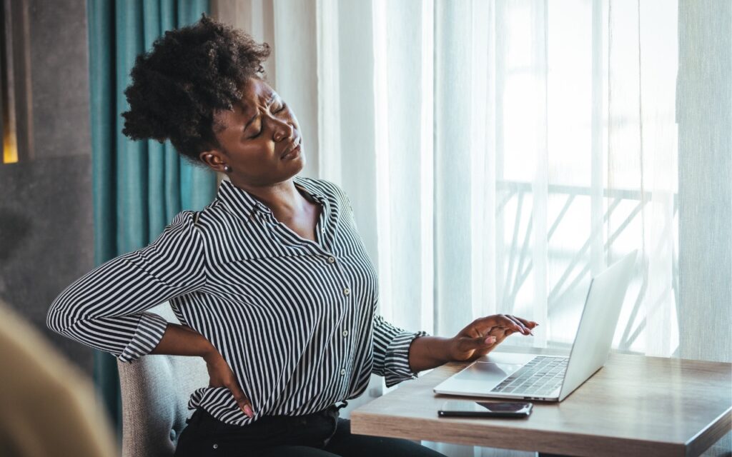 A woman sitting at a desk with a laptop holds her lower back and winces in pain, suggesting discomfort or back pain—highlighting the need for advice for good office ergonomics to prevent issues from poor posture or prolonged sitting.