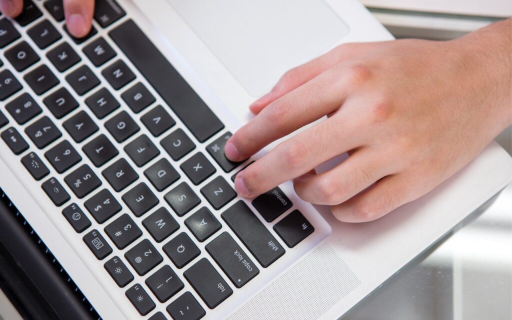 A close-up of a person's hands typing on a white laptop with black keys. The image emphasizes working on a computer, suggesting shortcuts like Press Control-Return in Sequoia to Open Contextual Menus while typing.