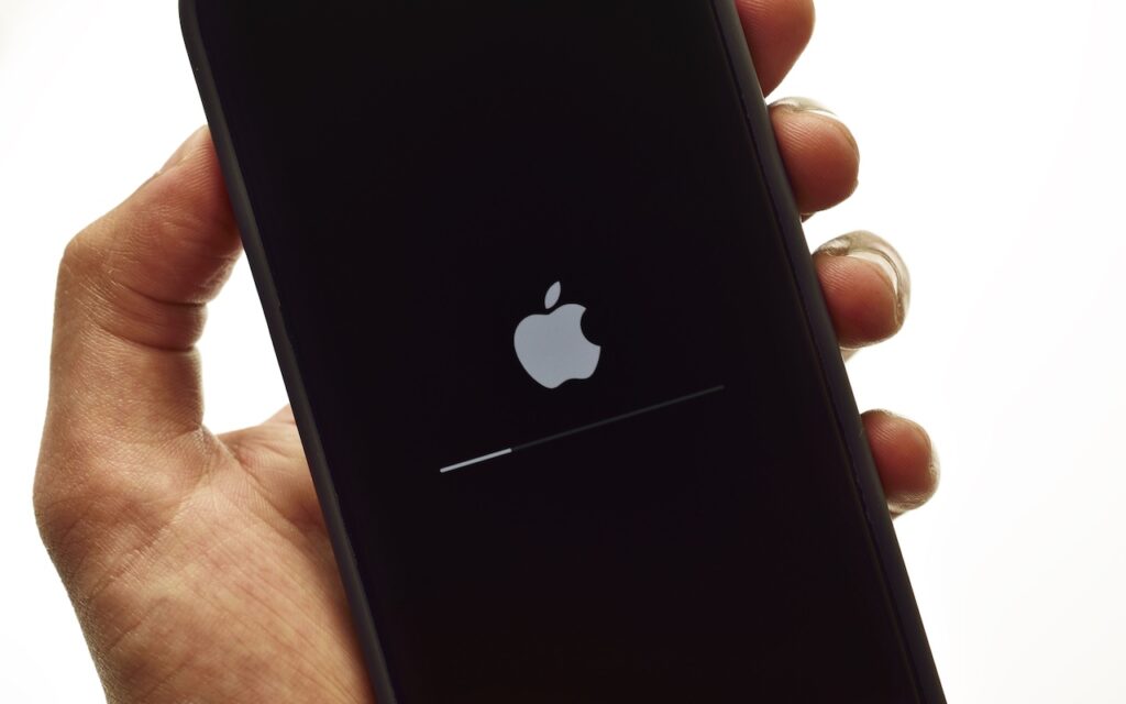 A hand holding a smartphone with an Apple logo and a loading bar displayed on the screen, suggesting the device is recovering from a failed update with a nearby iPhone or iPad.