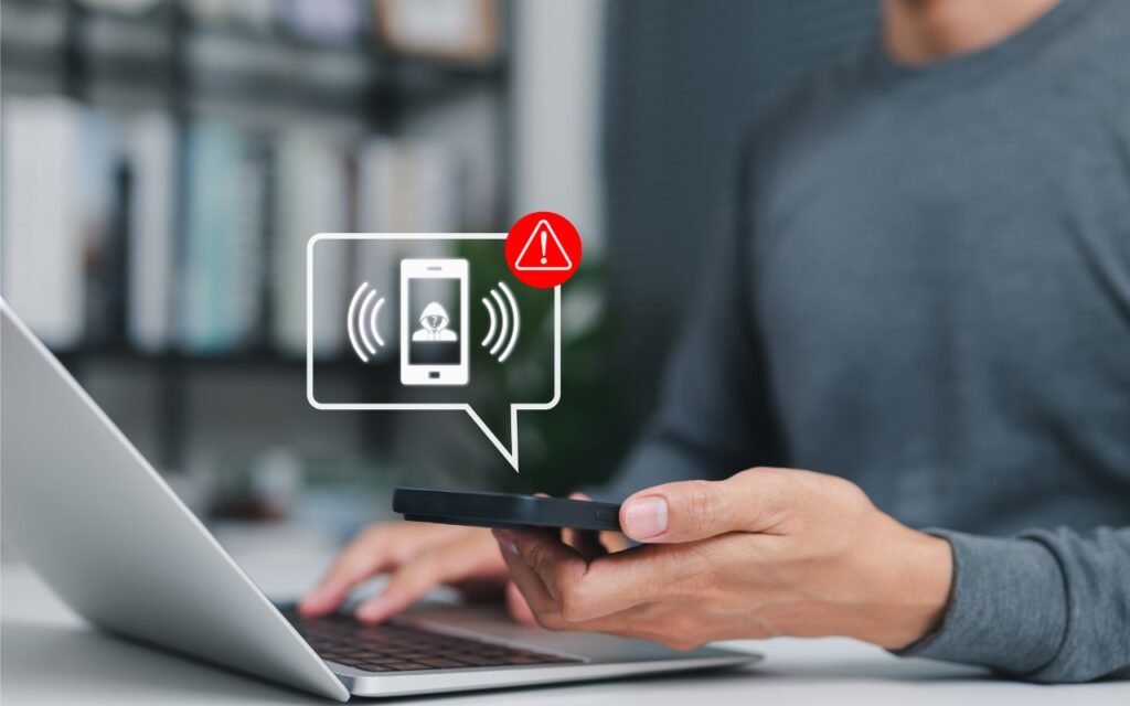 A person using a smartphone and laptop receives a warning notification on the phone, shown by a digital alert icon and exclamation mark. The scene suggests a security or system alert—reminding users to watch out for modern tech support scams.