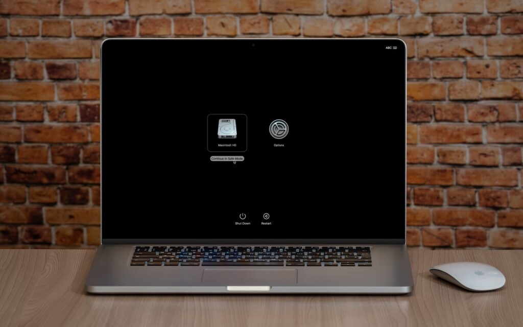 A MacBook on a wooden desk displays its startup menu with options for Macintosh HD, Options, and What Is Safe Mode? A mouse sits to the right of the laptop, set against a brick wall in the background.