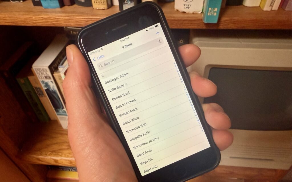A hand holds a smartphone displaying contacts in the iCloud section with names in alphabetical order, illustrating How to Display Contacts with Last Names First. A bookshelf filled with books is visible in the background.