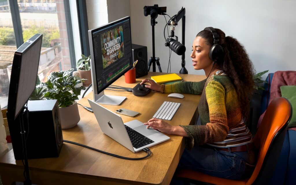 A woman wearing headphones speaks into a microphone while using a computer at a desk with a monitor, laptop—possibly the new MacBook Pro with Apple M5 Chip—keyboard, camera, and plants in a bright home office setup.