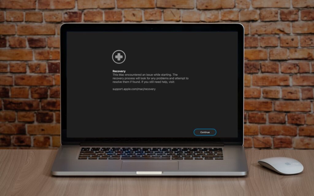 A MacBook displaying a Recovery screen referencing macOS 26 Tahoe Introduces New Recovery Assistant, placed on a wooden desk beside a white Apple Magic Mouse, with a brick wall background.
