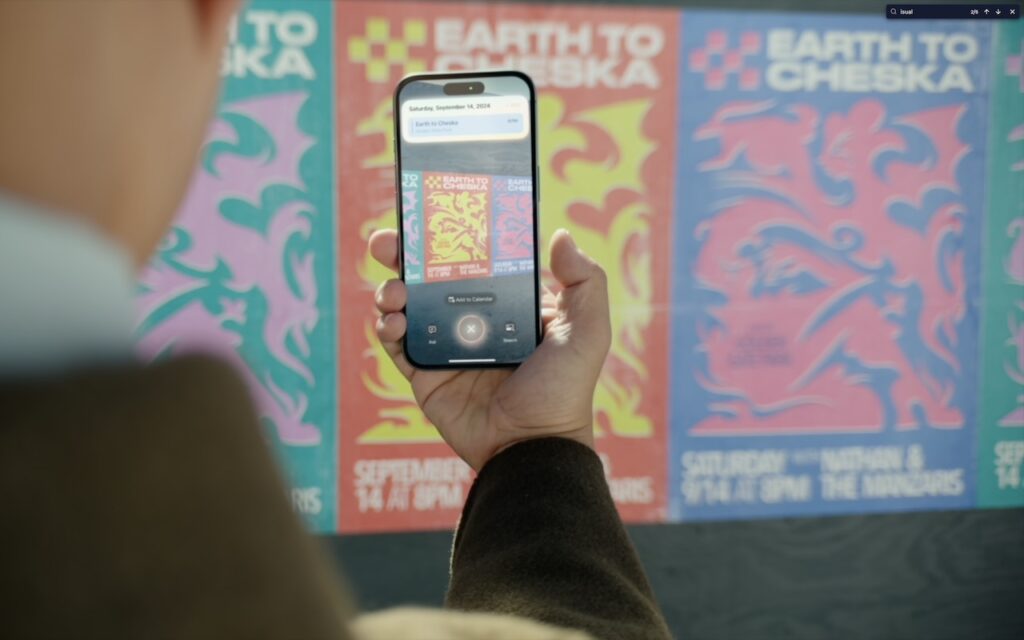 A person holds a smartphone up to colorful Earth to Cheska event posters on a wall, using the phone’s camera to scan and identify details—demonstrating how to Make the Most of Visual Intelligence on the iPhone.