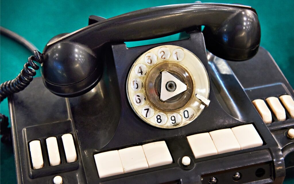 Close-up of a vintage black rotary telephone with a circular dial and white number buttons, resting on a green surface—like something you’d see in the retro redesign of iOS 26’s Phone App. The handset sits on top, with several white keys visible.