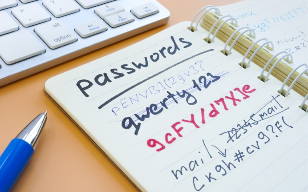 An open notebook labeled passwords lists various passwords, some crossed out in red. A blue pen and part of a keyboard are visible nearby—much like what you might see in the new Updated Passwords App Adds History feature.