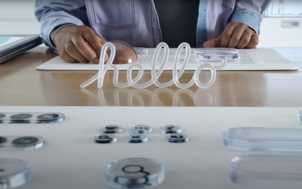 A person in a lab coat stands behind a desk with lab equipment and clear objects, including a transparent sign that spells hello in cursive letters.