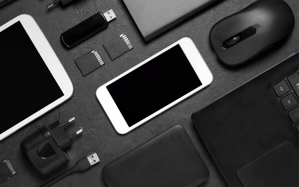 A flat lay of various tech gadgets and accessories on a dark surface, showcasing A Few of Our Favorite Things: 2025 Tech Edition—featuring a smartphone, tablet, laptop, mouse, USB flash drives, SD cards, power adapters, and charging cables.