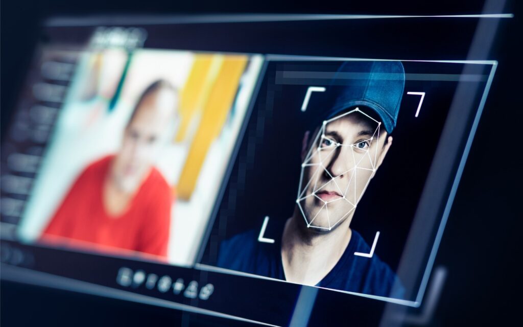 A digital screen displays facial recognition software analyzing a man in a blue cap, with facial mapping lines visible, urging viewers to Be Alert for Deepfake Phishing Scams alongside a blurred image of another person in a red shirt.