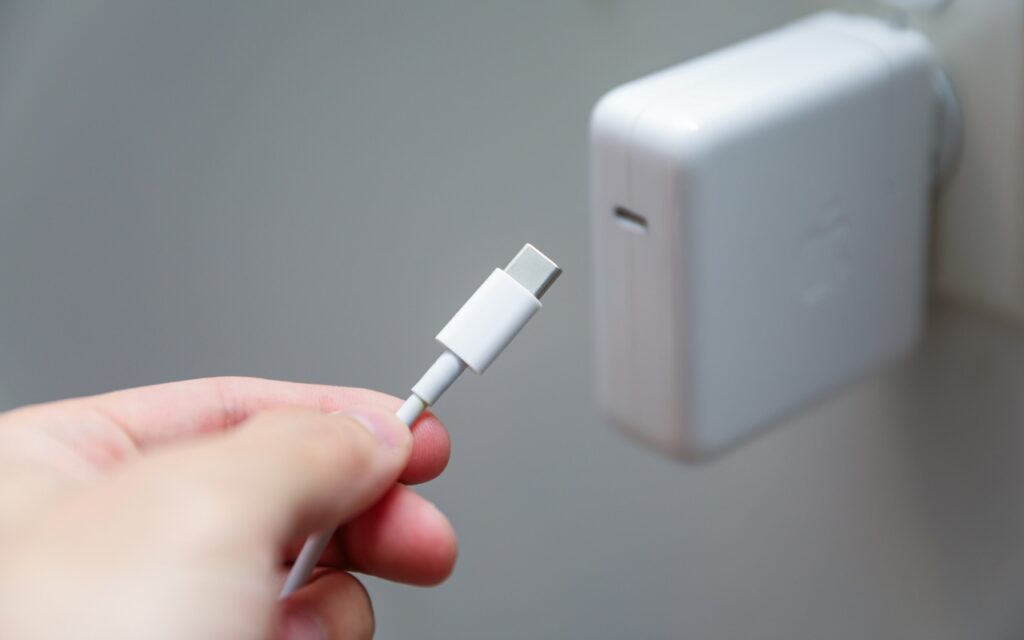 A hand holds a USB-C charging cable near a white wall charger plugged into a wall outlet—Charge Your MacBook with the Right Power Adapter for efficient and reliable performance.