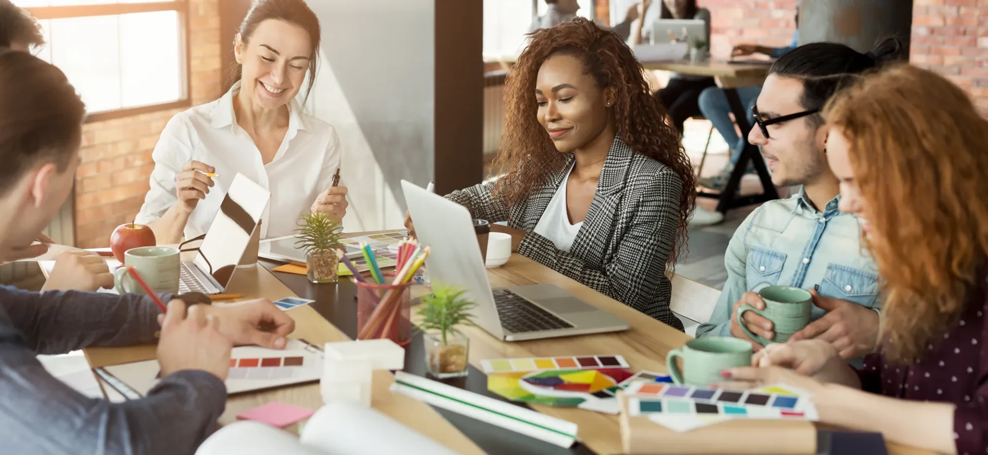 A group of five people sit around a table with laptops, notebooks, coffee mugs, and color swatches, collaborating in a modern, well-lit office space with large windows.