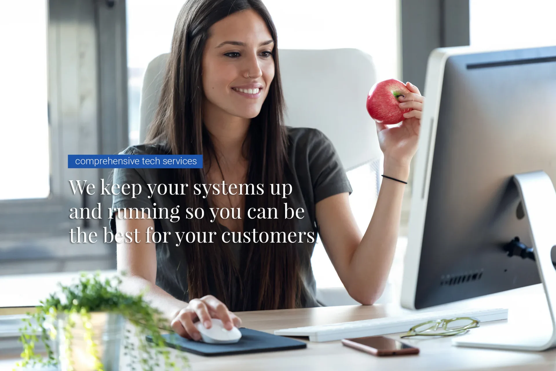 A woman smiles at her computer while holding an apple. Text on the image reads, &ldquo;We keep your systems up and running so you can be the best for your customers.&rdquo;.