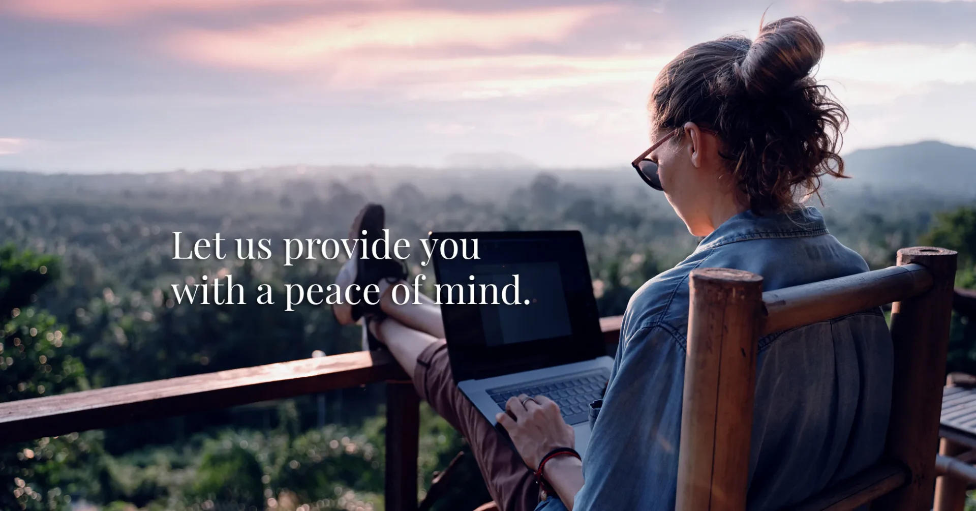 A person with sunglasses works on a laptop outdoors on a balcony at sunset, overlooking greenery. Text reads, &ldquo;Let us provide you with a peace of mind.&rdquo;.